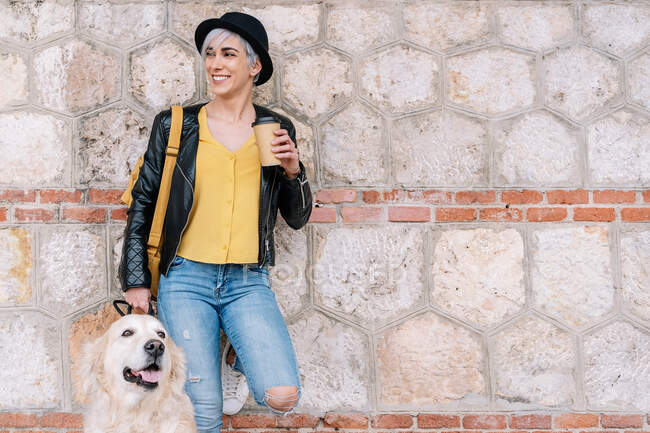 Young stylish female millennial in leather jacket and hat drinking coffee while standing looking away in city street stone wall with cute Golden Retriever dog on leash — Stock Photo