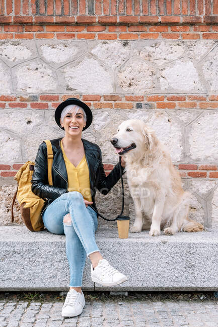 Trendy happy young female owner with dyed hair in stylish leather jacket and hat with adorable Golden Retriever dog while sitting on stone bench on street — Stock Photo