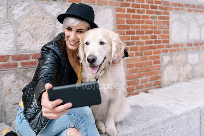 Happy young lady in trendy leather jacket and hat smiling while taking selfie with cute Golden Retriever dog sitting near brick building on street — Stock Photo