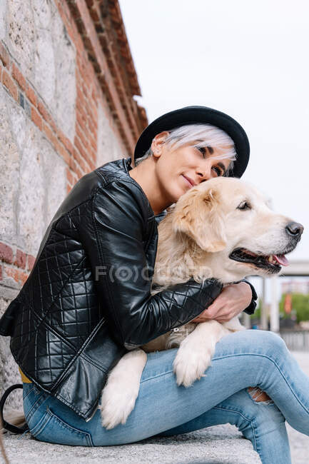 From below trendy young female owner with dyed hair in stylish leather jacket and hat hugging adorable Golden Retriever dog while sitting on stone bench on street — Stock Photo