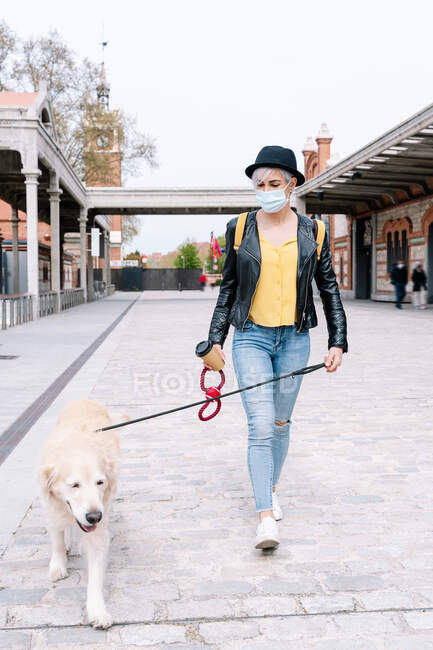 Full length of young stylish female millennial in leather jacket and medical mask walking in city park with cute Golden Retriever dog on leash — Stock Photo