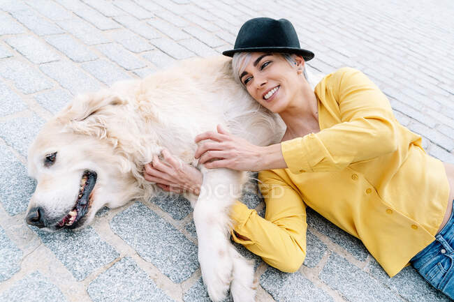 Smiling young female with felt hat lying down on charming purebred dog on pavement in daylight — Stock Photo