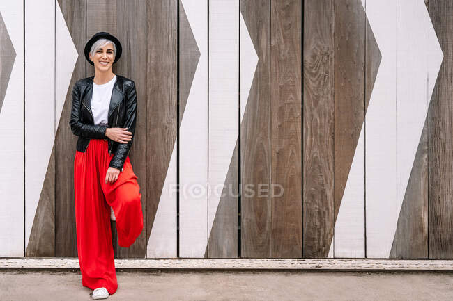 Mulher alegre em roupas da moda em pé na passarela enquanto olha para a câmera contra cerca de madeira — Fotografia de Stock