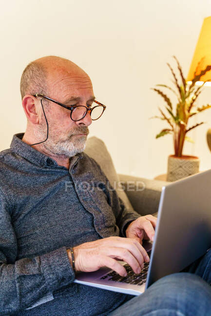 Hombre mayor en gafas de navegación por Internet en el ordenador portátil mientras está sentado en el sofá en casa - foto de stock