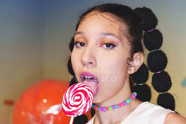 Stylish African American female licking sweet lollipop and looking at camera on background of glowing neon illumination in studio — Stock Photo