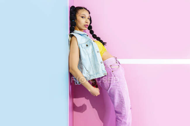 Side view stylish cool African American female in fashionable outfit standing in corner of studio with pastel color walls and looking at camera — Stock Photo