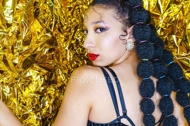 Вид сбоку на модную афроамериканку в блестящем наряде, стоящую на фоне золотой фольги в студии и смотрящую в камеру — стоковое фото