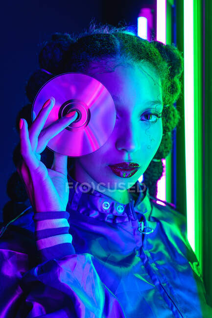 Stilvolle Afroamerikanerin in Vintage-Kleidung steht mit Compact Disc im dunklen Studio mit leuchtender Neonbeleuchtung und blickt in die Kamera — Stockfoto