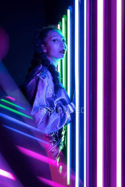 Side view of stylish African American female looking at camera in dark studio with colorful glowing neon lights — Stock Photo