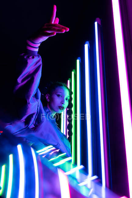 Seitenansicht einer stilvollen Afroamerikanerin, die in einem dunklen Studio mit bunten leuchtenden Neonlichtern in die Kamera blickt — Stockfoto