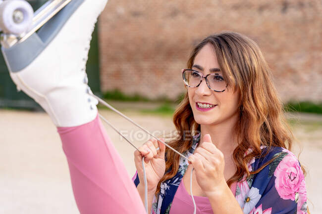 Молодая женщина в очках завязывает шнурки роликового конька, сидя с поднятой ногой на улице — стоковое фото
