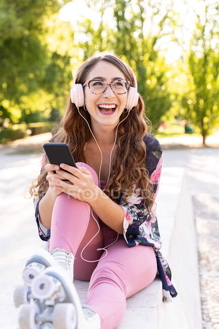 Весела молода жінка в окулярах і роликах переглядає інтернет на мобільному телефоні, слухаючи музику з навушників. — Stock Photo