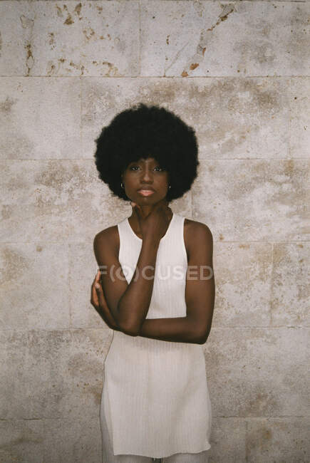 Self assured young black female model with dark Afro hair in white top touching neck and looking at camera against tiled wall — Stock Photo