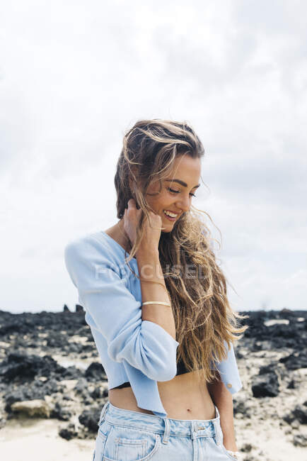 Elegante viajera femenina feliz de pie con los ojos cerrados solo en medio de un terreno desierto con piedras volcánicas bajo el cielo azul nublado mientras disfruta de la aventura de verano en la isla de Lanzarote en España - foto de stock