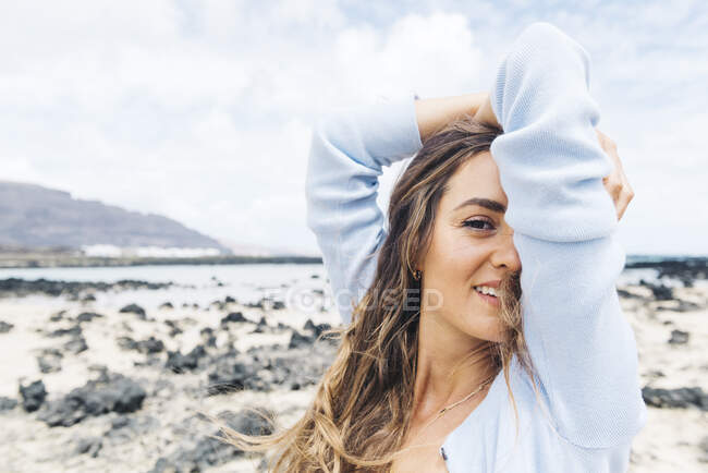 Stylish happy female traveler standing looking at camera amidst deserted terrain with volcanic stones under blue cloudy sky while enjoying summer adventure on Lanzarote island in Spain — Stock Photo