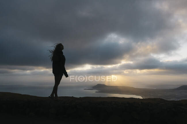 Vista lateral de silueta de mujer viajera irreconocible de pie en la colina con los brazos extendidos en las tierras altas y disfrutando de la puesta de sol en Lanzarote - foto de stock