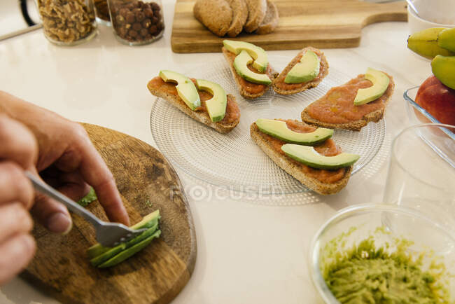 З висоти анонімний чоловік кладе шматочки свіжого авокадо на хліб з соусом під час приготування здорового сніданку. — стокове фото