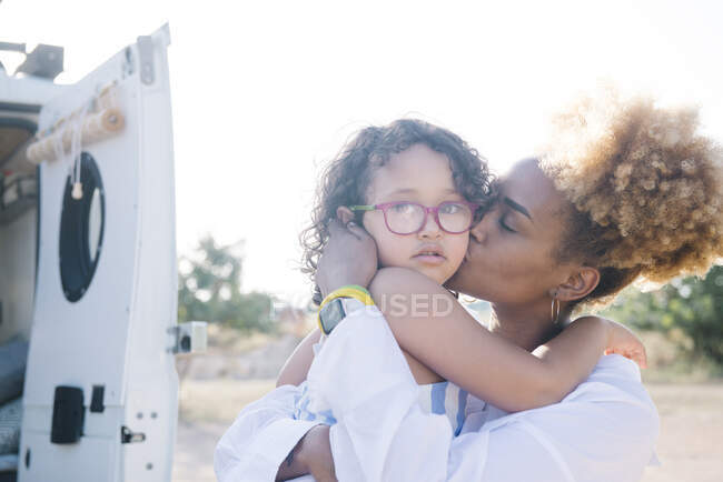 Side view of loving African American mother gently kissing cute daughter in cheek while standing near parked camper during trip in nature — Stock Photo
