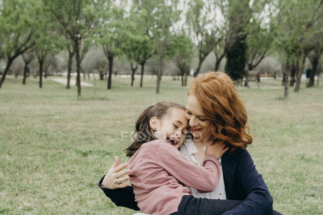 Cheerful redhead mother hugging cute laughing preschooler daughter while having fun together in green park — Stock Photo