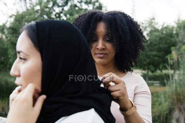 Mujer afroamericana ajustando el velo de la amiga musulmana mientras está de pie en el parque - foto de stock