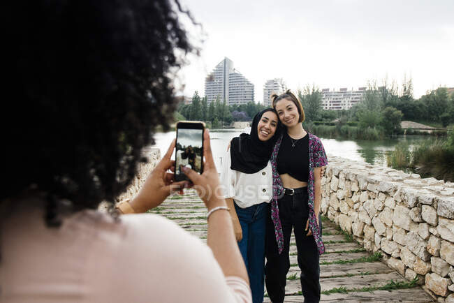 Mulher irreconhecível tirar foto de amigos femininos multirraciais em pé perto do lago na cidade — Fotografia de Stock