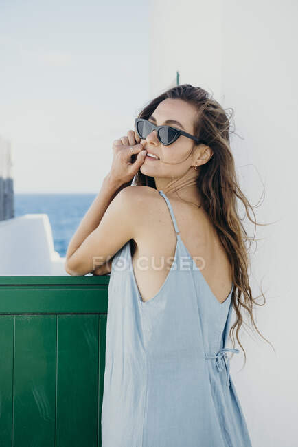 Side view of young barefoot female in sundress and sunglasses leaning against green gate of a house looking at camera while relaxing alone in summertime near sea — Stock Photo