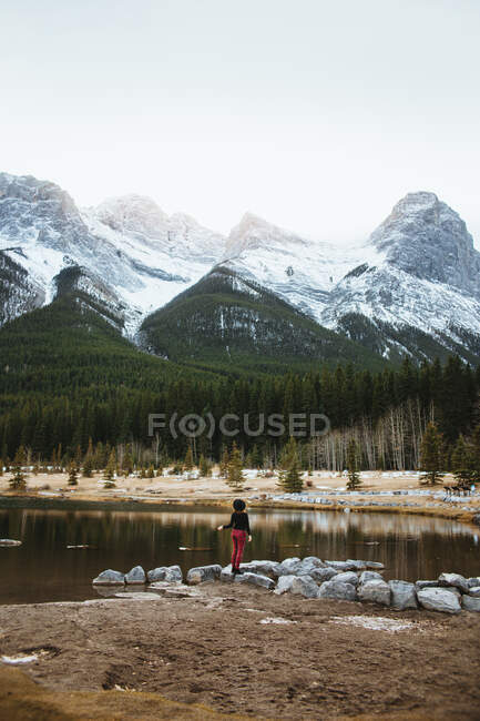 Обратный вид на неузнаваемую женщину-туристку, стоящую в одиночестве на берегу Куорри-Лейк против величественных лесистых гор со снежными скалистыми вершинами в Национальном парке Банф в Канаде — стоковое фото