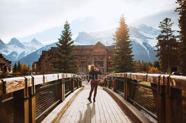 Rückansicht einer unkenntlichen Reisenden auf einer hölzernen Fußgängerbrücke vor einer bergigen Landschaft mit schneebedeckten Gipfeln, während sie Herbstferien in der Stadt Canmore in der Nähe des Banff-Nationalparks in Kanada verbringt — Stockfoto