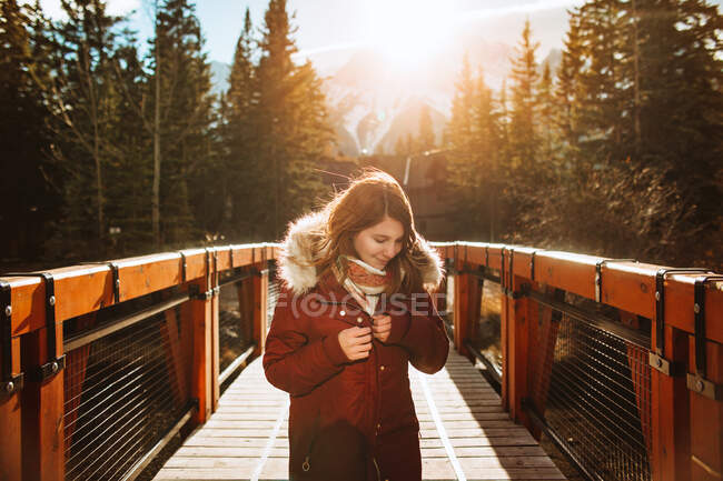 Giovane viaggiatrice in capispalla godendo soleggiata giornata autunnale mentre in piedi sul ponte di legno sul fiume contro alberi di conifere e montagne nel Banff National Park in Canada — Foto stock