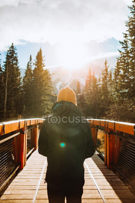 Обратный вид на неузнаваемого мужчину-путешественника на деревянном пешеходном мосту против горного ландшафта с заснеженными вершинами во время осеннего отпуска в городе Канмор возле национального парка Банф в Канаде — стоковое фото