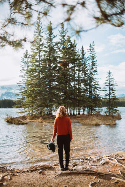 Обратный вид на неузнаваемую женщину, стоящую, любуясь зелеными хвойными деревьями, растущими на островке посреди озера Два Джек против облачно-голубого неба в Альберте, Канада — стоковое фото