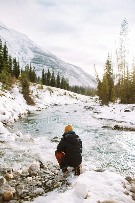 Back view of anonymous hiker sitting on snowy coast of rapid brook on winter day in Marble Canyon, British Columbia — Stock Photo