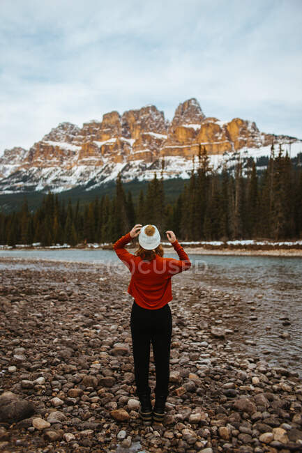 Back view of anonymous female traveler admiring snowy Castle Mountain and coniferous trees while standing on river coast in Banff National Park — Stock Photo