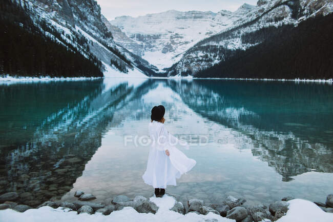 Обратный вид анонимной женщины в белом платье, стоящей в сторону чистой воды озера Луиза против снежного горного хребта в зимний день в Альберте, Канада — стоковое фото