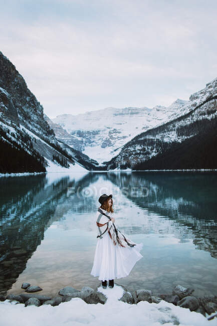 Вид сбоку женщины в белом платье и шарфе, стоящей к чистой воде озера Луиза на фоне заснеженного горного хребта в зимний день в Альберте, Канада — стоковое фото