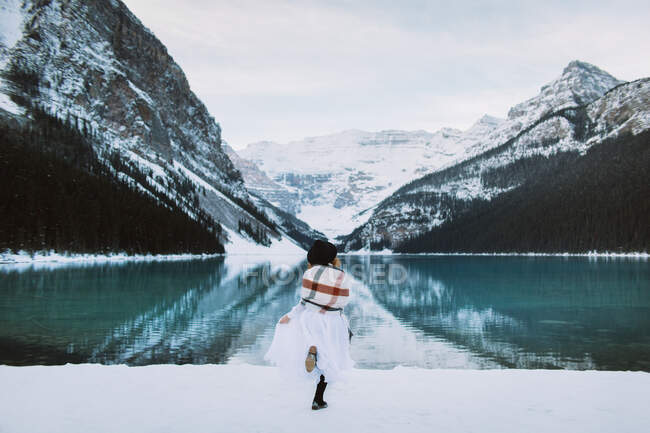 Rückansicht einer anonymen Frau in weißem Kleid und Schal, die an einem Wintertag in Alberta, Kanada, gegen den schneebedeckten Bergrücken auf das saubere Wasser des Lake Louise zuläuft — Stockfoto