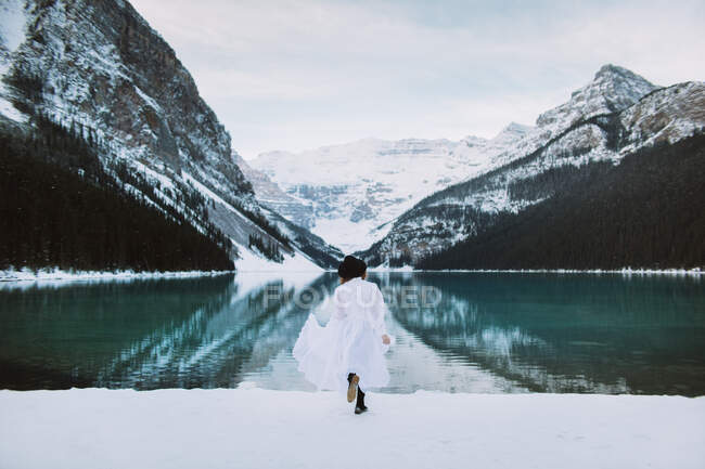 Rückansicht einer anonymen Frau im weißen Kleid, die an einem Wintertag in Alberta, Kanada, gegen den schneebedeckten Bergrücken in Richtung des sauberen Wassers des Lake Louise rennt — Stockfoto