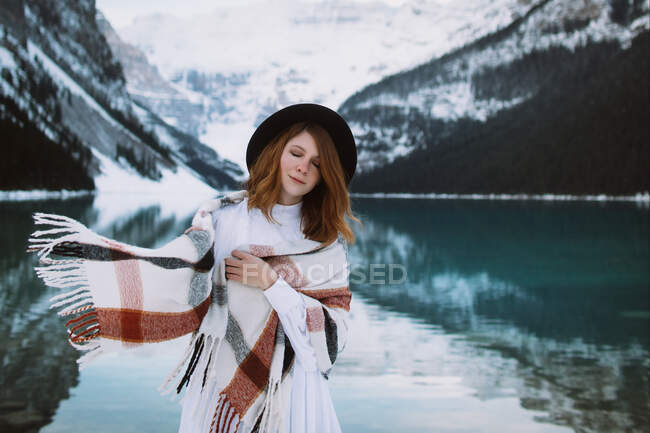 Female in white dress and scarf standing with eyes closed near clean water of lake Louise against snowy mountain ridge on winter day in Alberta, Canada — Stock Photo