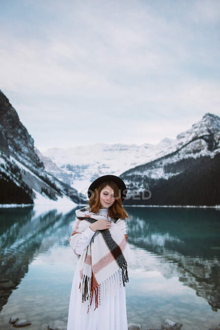 Weibchen in weißem Kleid und Schal stehen mit geschlossenen Augen in der Nähe des klaren Wassers des Lake Louise gegen den schneebedeckten Bergrücken an einem Wintertag in Alberta, Kanada — Stockfoto