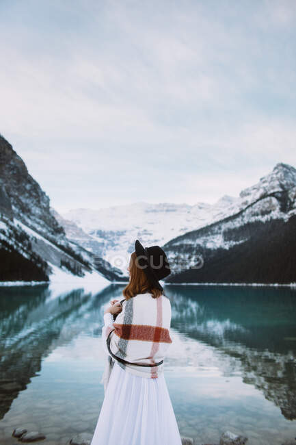 Visão traseira da fêmea anônima em vestido branco e cachecol em pé em direção à água limpa do lago Louise contra cume de montanha nevada no dia de inverno em Alberta, Canadá — Fotografia de Stock