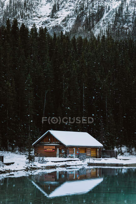 Деревянная хижина расположена на снежном берегу спокойного озера Луиза возле хвойных лесов и горных хребтов в холодный зимний день в Национальном парке Банф — стоковое фото
