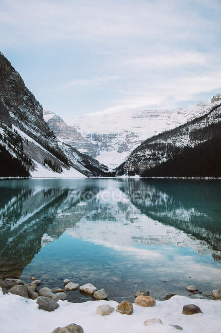 Eau propre du paisible lac Louise reflétant la crête enneigée des montagnes et le ciel nuageux lors de la journée d'hiver en Alberta, Canada — Photo de stock