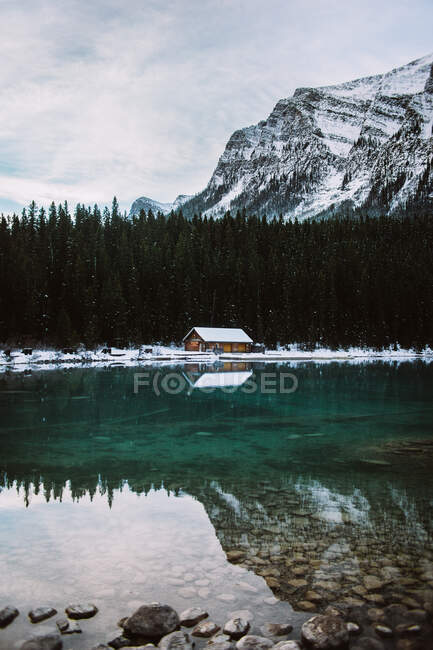 Cabane en bois située sur la côte calme du lac Louise près de la forêt de conifères et de la montagne enneigée lors de la journée d'hiver en Alberta, Canada — Photo de stock