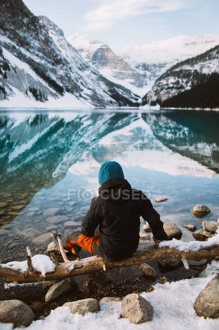 Visão traseira do turista anônimo em suéter e chapéu levantando braços e alongamento enquanto sentado na costa do Lago Louise contra cume de montanha nevada no dia de inverno em Alberta, Canadá — Fotografia de Stock