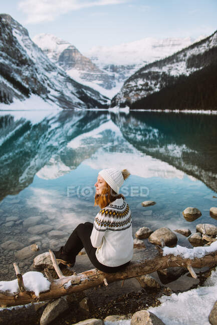 Back view of thoughtful female tourist in sweater and hat sitting with eyes closed on coast of Lake Louise against snowy mountain ridge on winter day in Alberta, Canada — Stock Photo