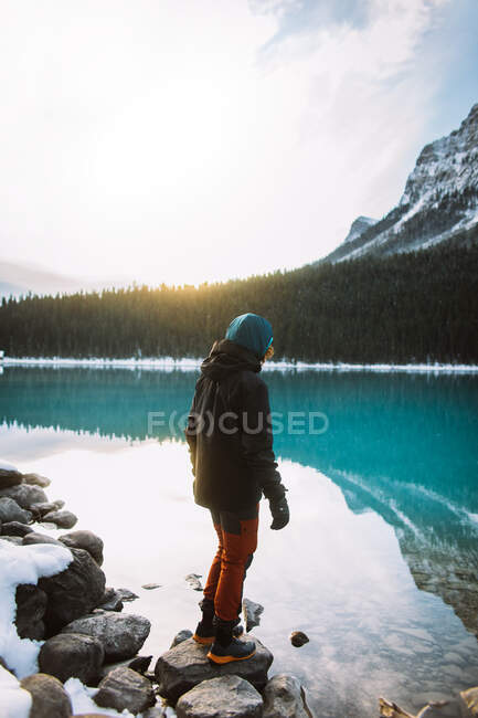 Viajero anónimo de cuerpo completo en ropa de abrigo de pie sobre rocas cerca de aguas tranquilas del lago Louise en la mañana en el Parque Nacional Banff - foto de stock