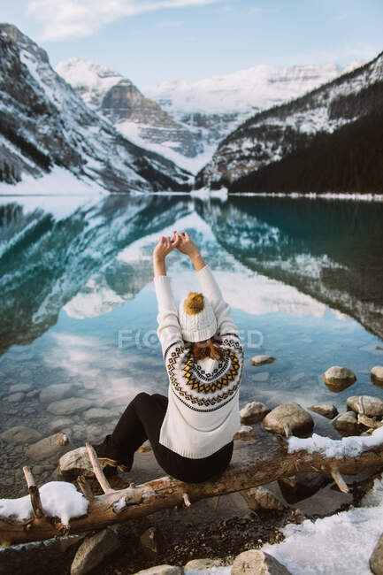Обратный вид анонимной туристки в свитере и шляпе, сидящей на берегу озера Луиза против снежного горного хребта в зимний день в Альберте, Канада — стоковое фото