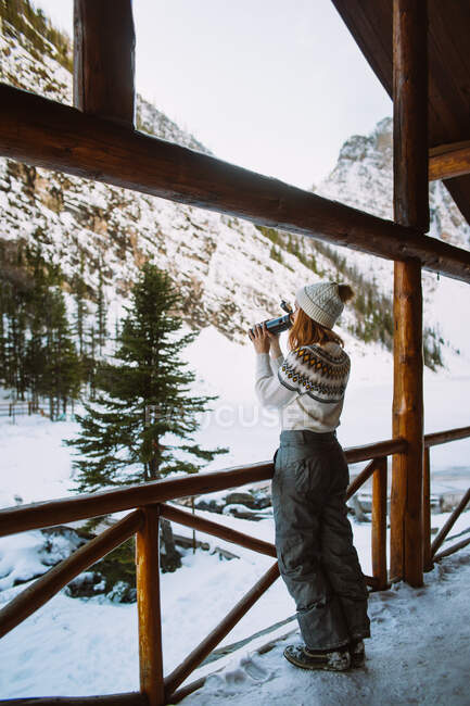 Viajante do sexo feminino desfrutando de bebida quente de garrafa térmica enquanto descansa em abrigo de madeira perto de montanhas nevadas no Parque Nacional Banff — Fotografia de Stock