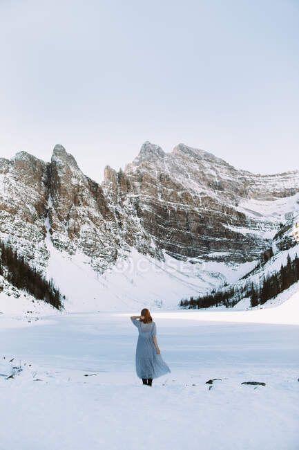 Rückansicht einer anonymen Frau im Kleid, die an einem Wintertag in Alberta, Kanada, in der Nähe des zugefrorenen Lake Louise vor einer verschneiten Bergkette steht — Stockfoto