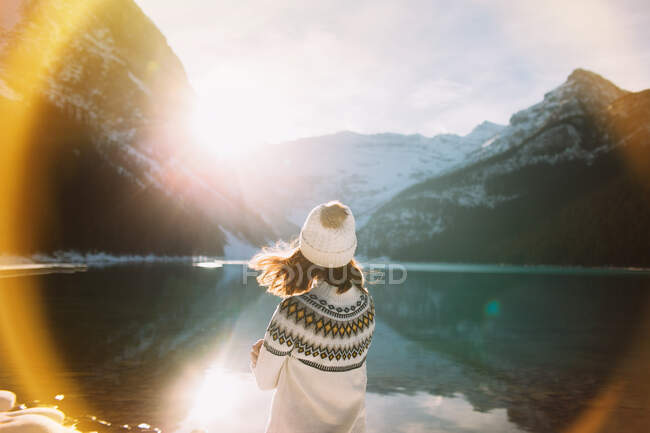 Задний вид анонимной женщины-туристки в теплой одежде, идущей против спокойного озера Луиза и стоящих гор в солнечное зимнее утро в Национальном парке Банф — стоковое фото